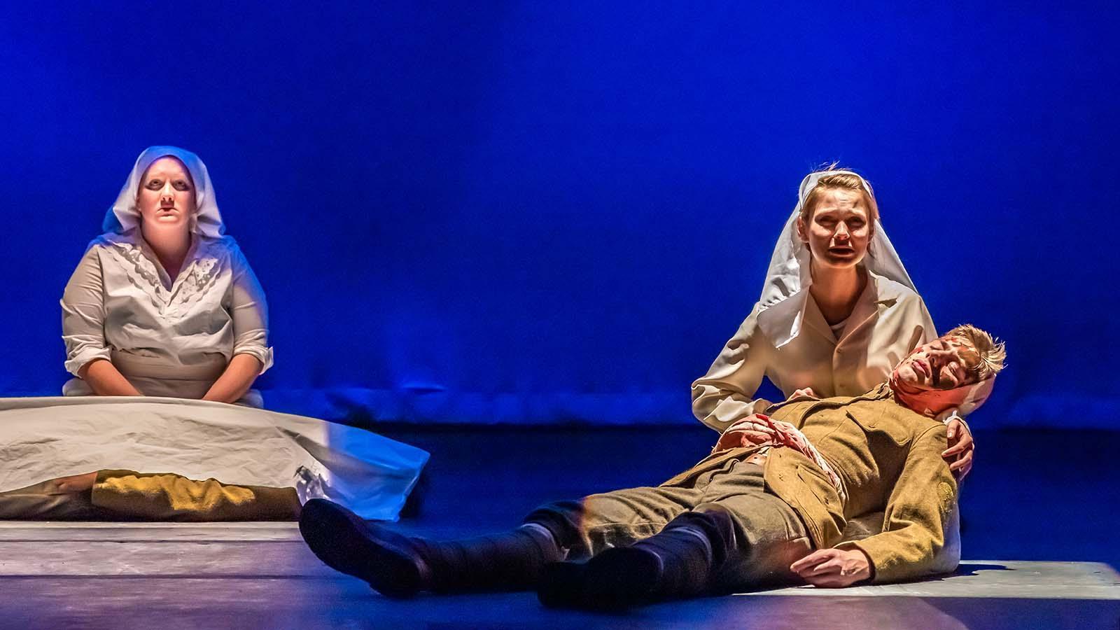 A student actress on stage as a WWI nurse on her knees holding an actor playing a fallen soldier. Another WWI nurse is seated behind them.